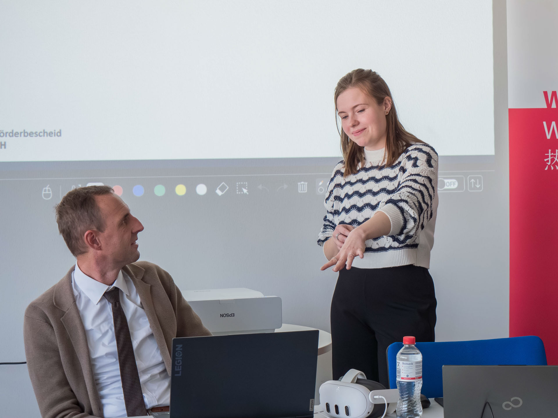 Eine Frau steht neben einem Mann, der an einem Tisch sitzt, und zeigt auf einen Bildschirm
